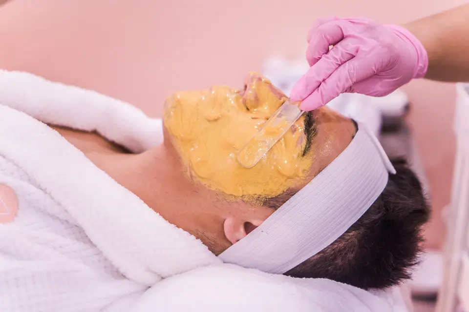 Esteticista con guantes aplicando una máscara facial con pincel a un cliente recostado.