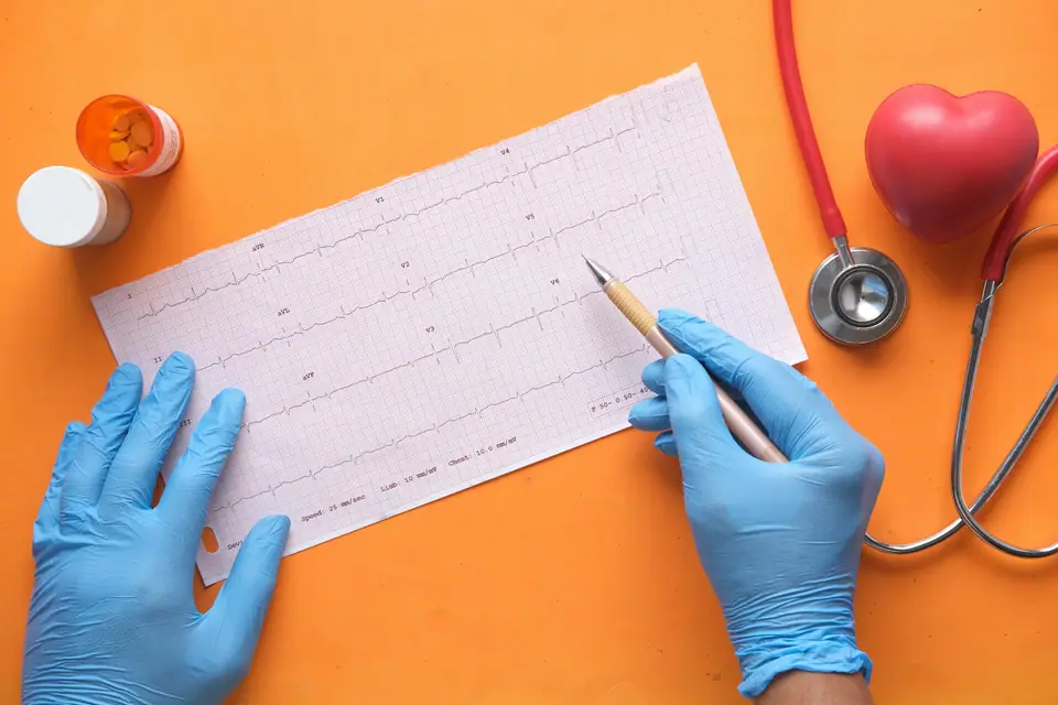 Manos con guantes señalando un trazado de electrocardiograma junto a un estetoscopio y un corazón de juguete.