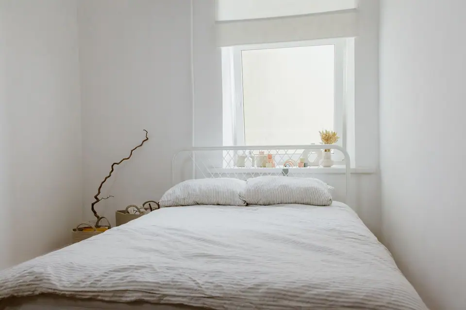 Dormitorio simple con cama de hierro blanca y ventana con luz natural.