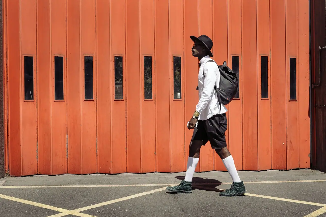 Hombre con sombrero, camisa blanca, bermuda negra y zapatillas caminando frente a un portón naranja