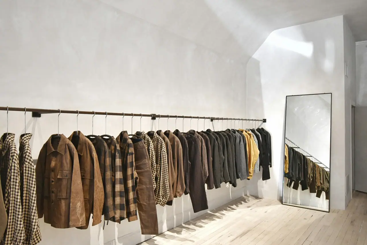 Interior minimalista de tienda con camisas y chaquetas colgadas en un barral y un espejo de pie