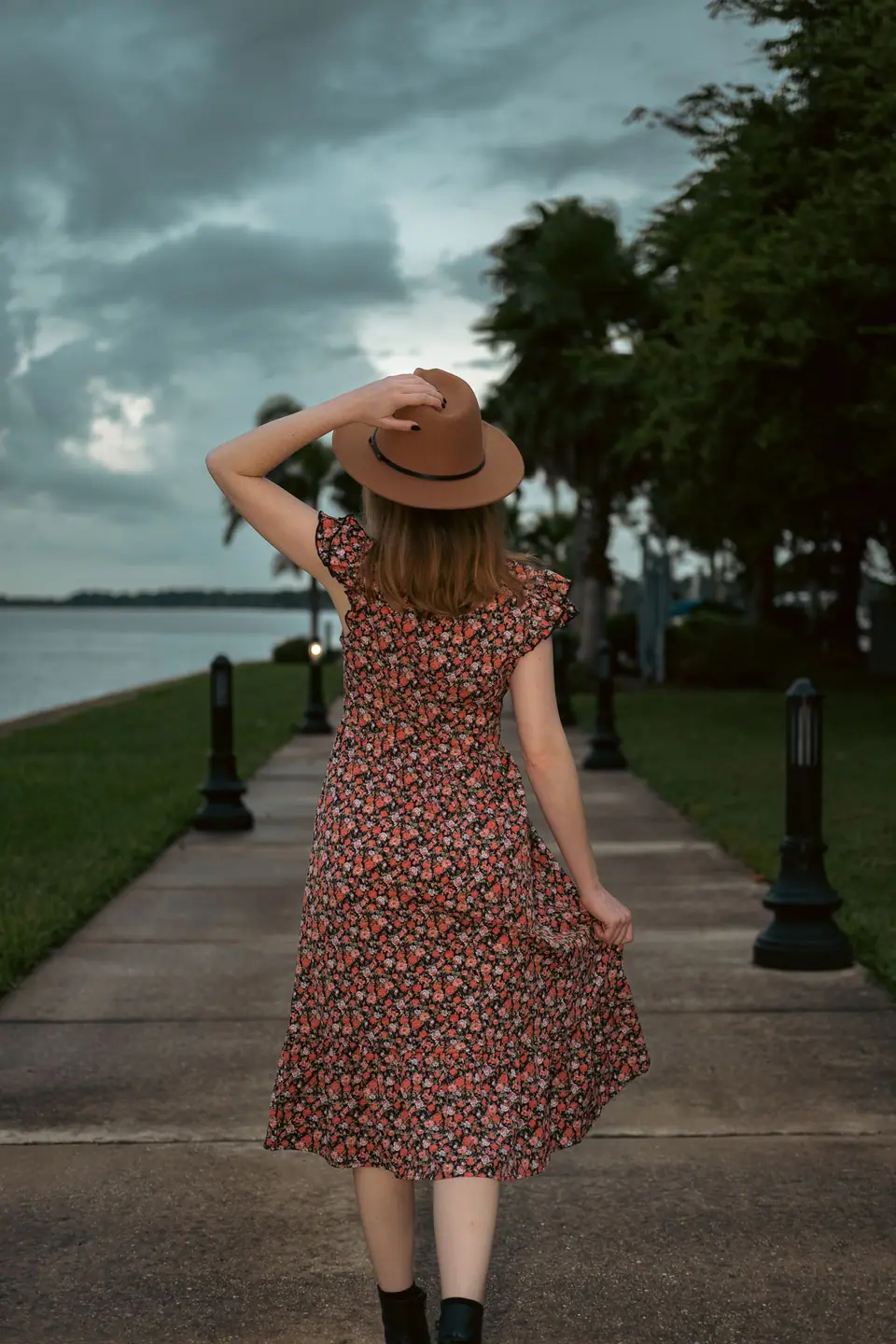 Mujer de espaldas con vestido midi floreado y sombrero caminando por un paseo junto al agua