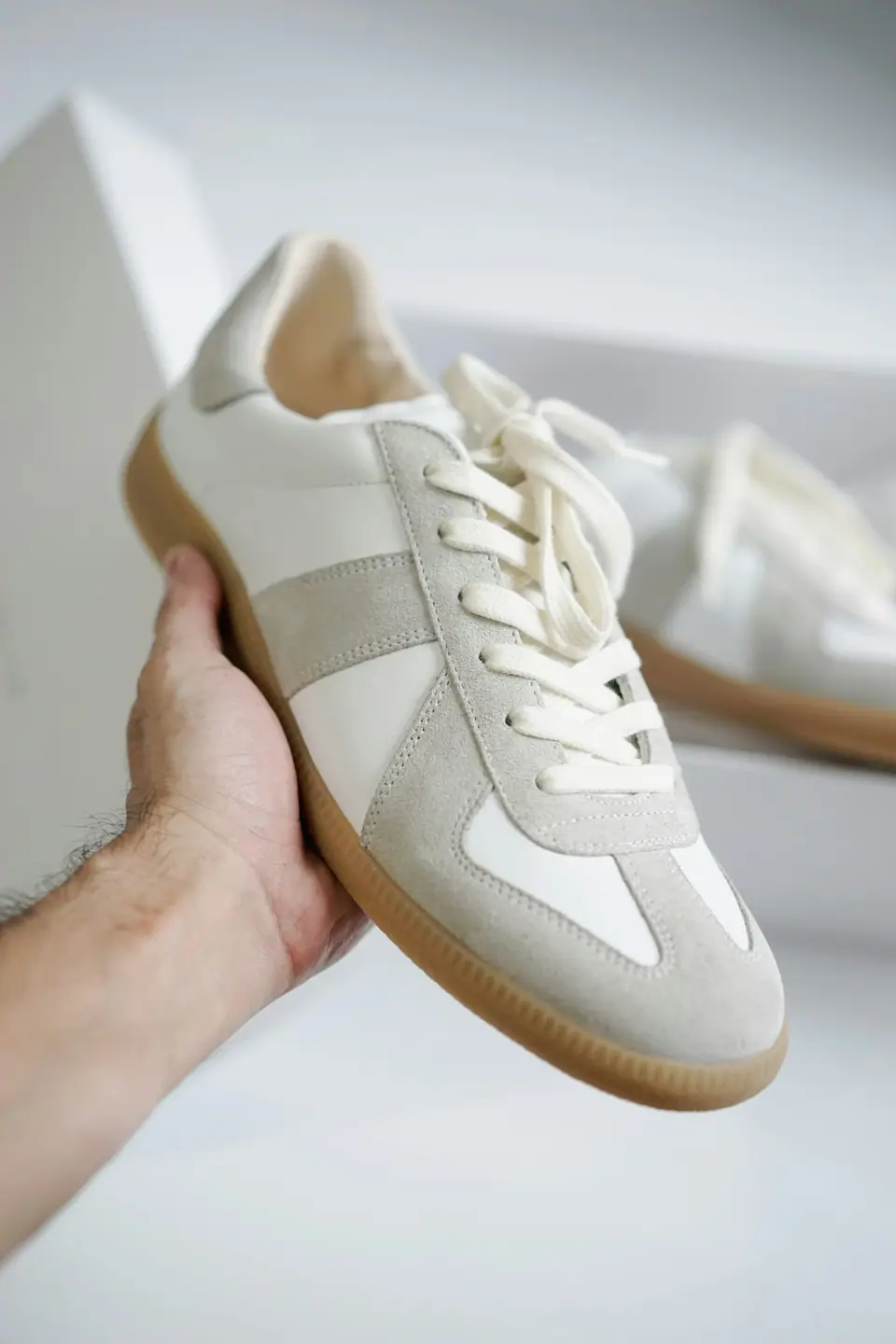 Zapatilla urbana blanca con detalles de gamuza gris y suela de goma, sostenida en la mano