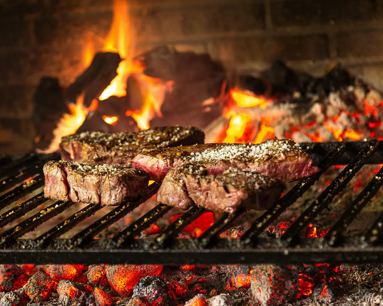 Cortes de carne con sal gruesa sobre la parrilla, con brasas encendidas y llamas de fondo