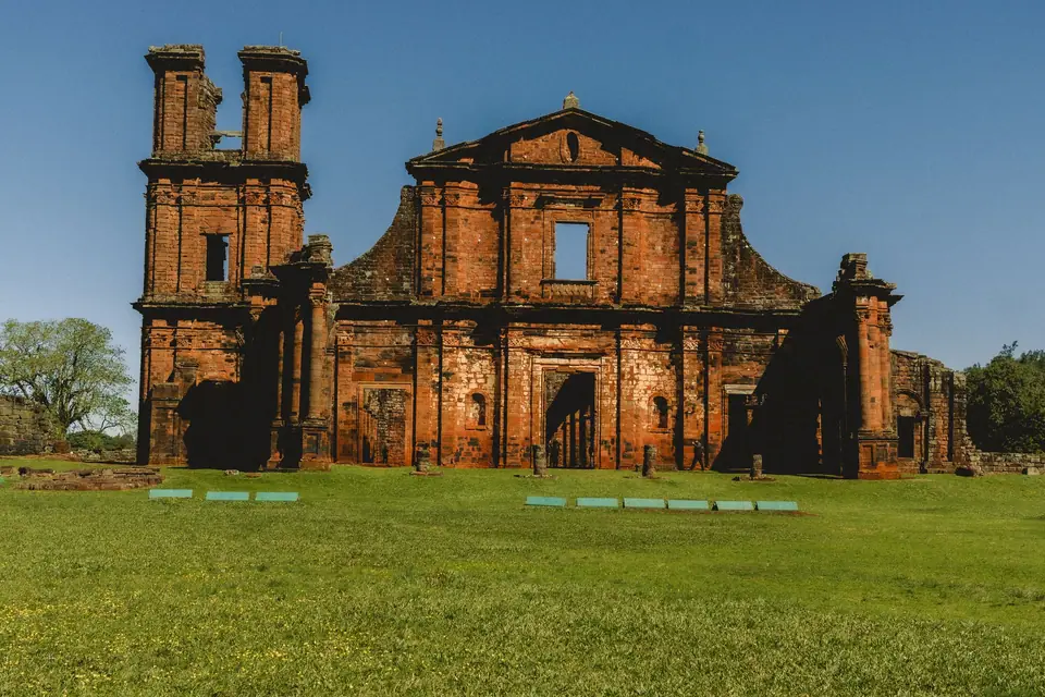 Ruinas de piedra rojiza de una iglesia de las misiones jesuíticas sobre un prado verde.